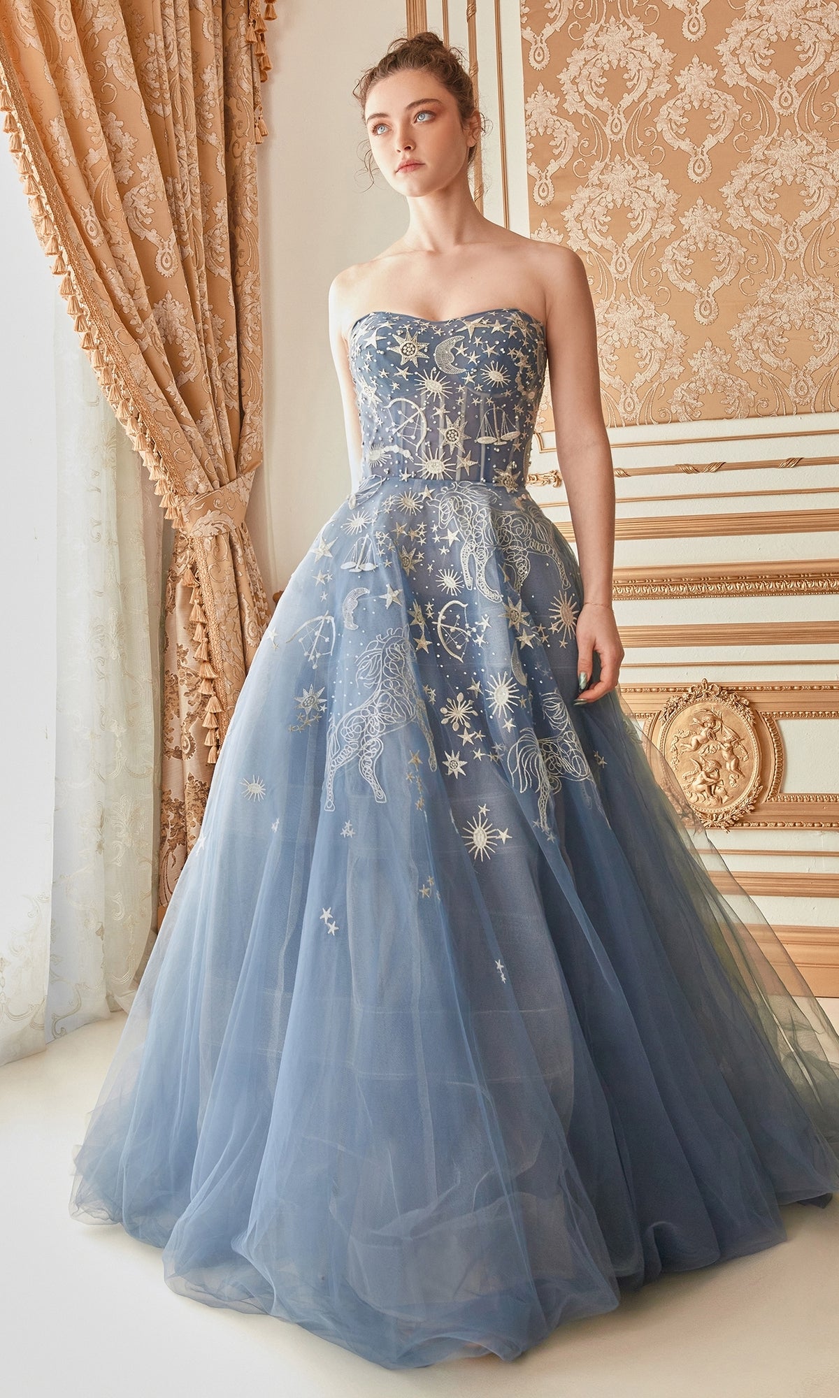 A woman stands in an ornate, vintage-style room adorned with gold accents and patterned curtains, wearing the Constellation-Print Strapless Prom Ball Gown A0890 featuring floor-length celestial embroidery.